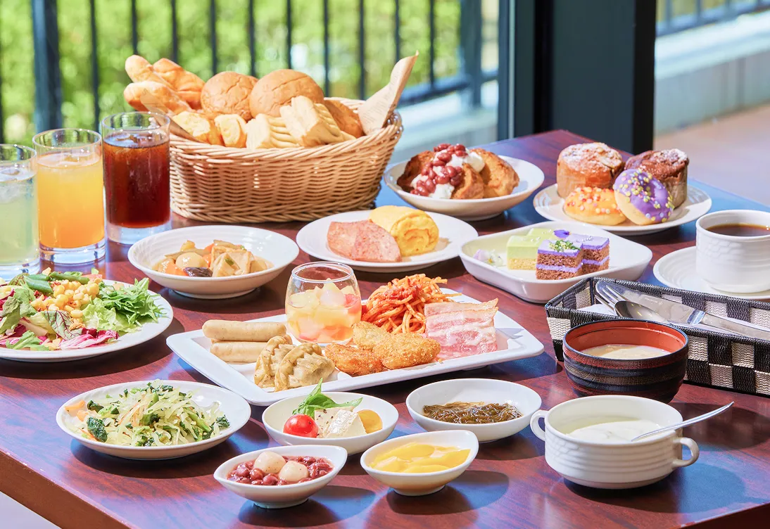 輝く青い海をバックに、テラス席で朝食を楽しむ女性。テーブルには色とりどりのフルーツやジュース、地元食材の料理がバラエティ豊かに並ぶ様子。