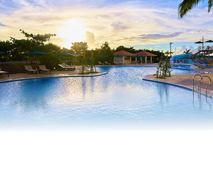 夕暮れ時の広々とした屋外プール。水面には夕日が反射し、周囲にはパラソルとデッキチェア、そしてヤシの木やウォータースライダーが見える開放的な風景。