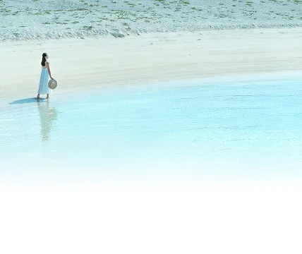 白いワンピースを着た女性が、透明度の高いエメラルドグリーンの海と白い砂浜の波打ち際を歩く、静かで美しい浜辺の景色。