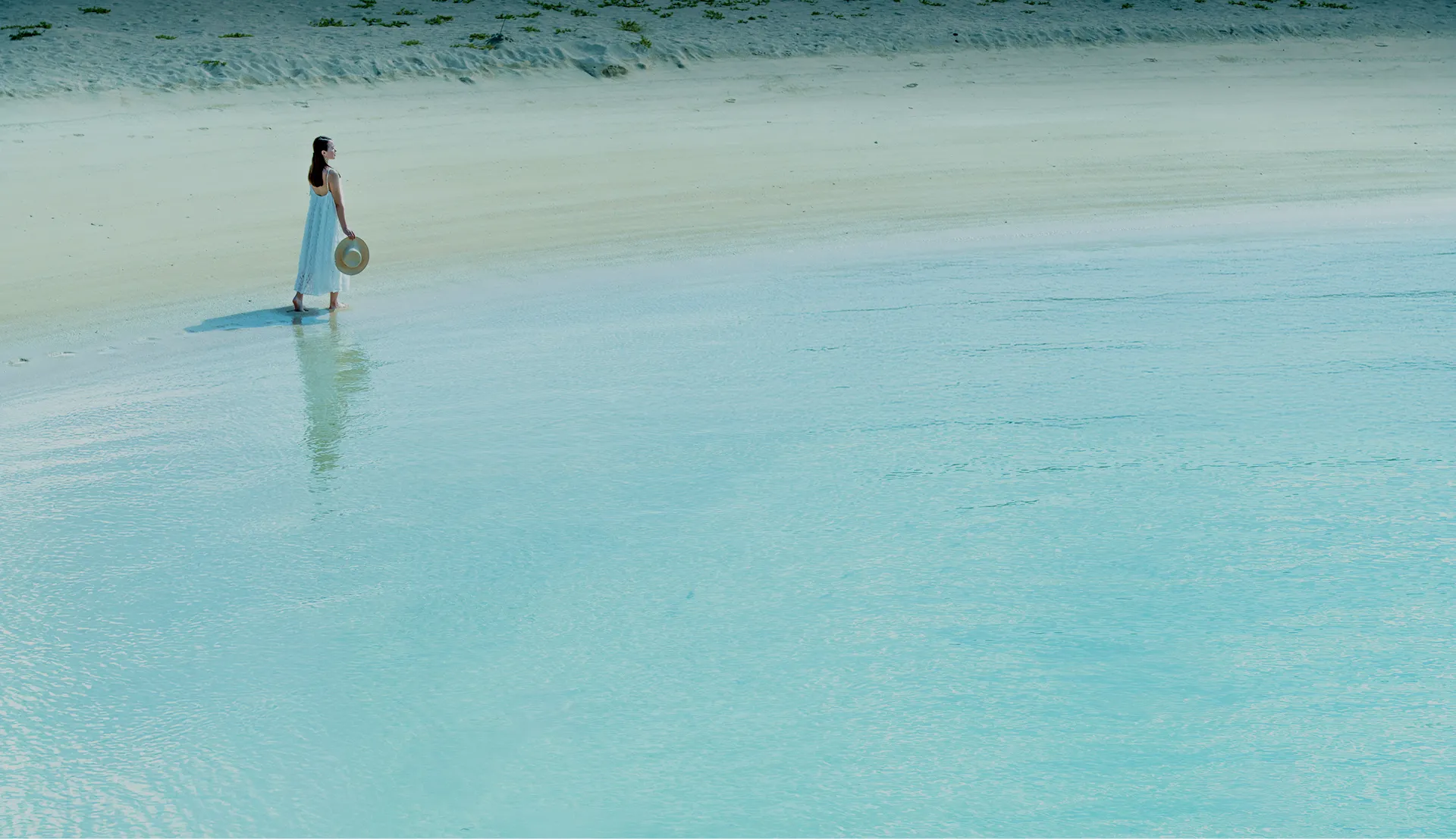 透き通るような美しい沖縄の海。白い砂浜と淡いブルーの海が溶け合う波打ち際を、麦わら帽子を手にした女性がゆったりと歩いています