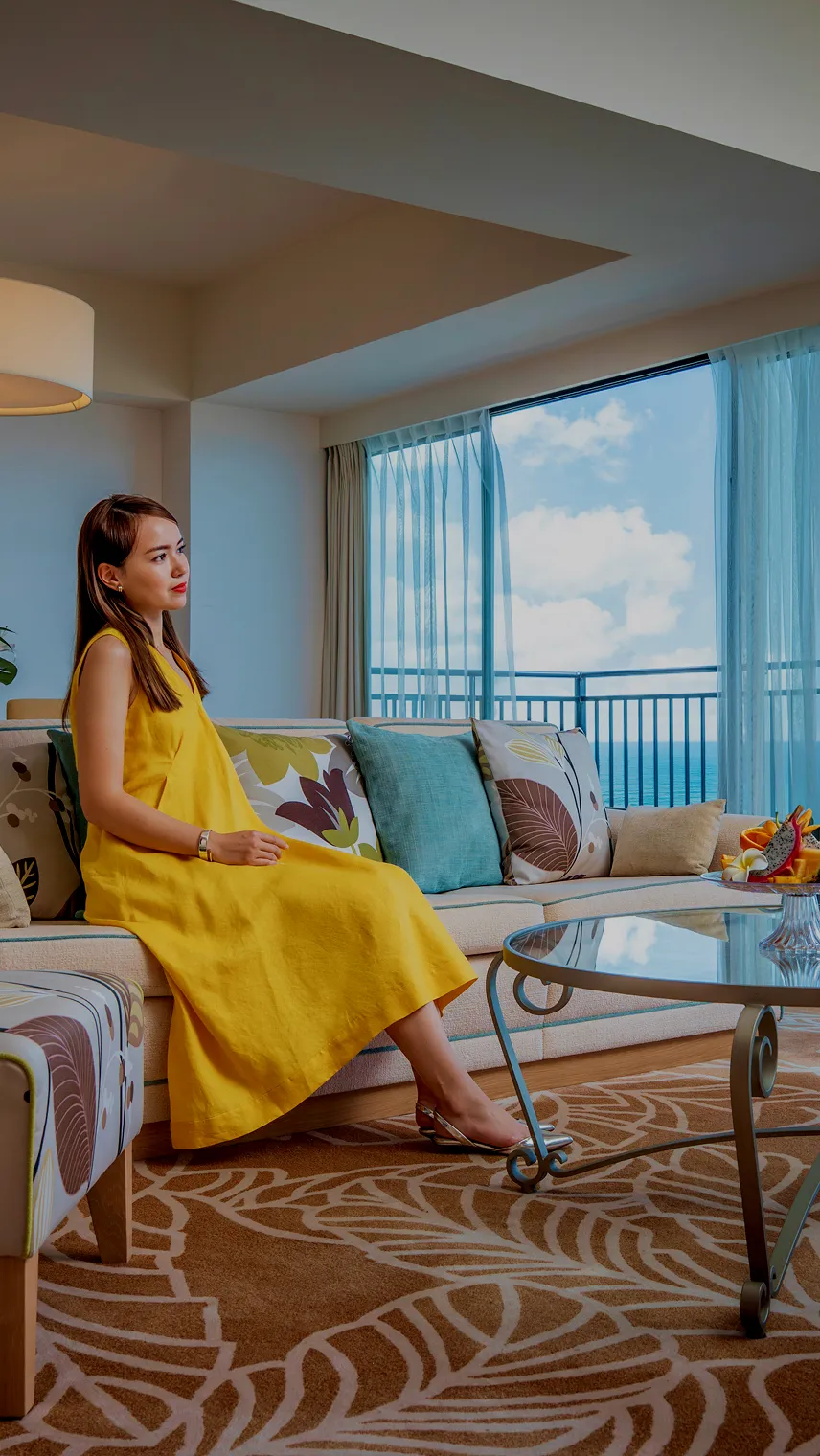 大きな窓一面にエメラルドグリーンの海と青空が広がる、明るく開放的な客室。落ち着いたインテリアの室内から、テラス越しに沖縄の絶景を一望できる様子