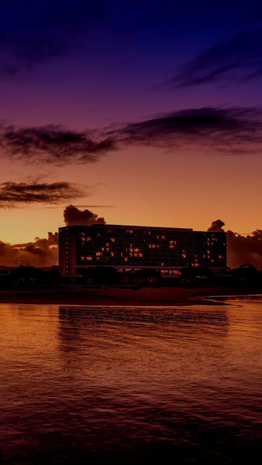刻一刻と表情を変える美しいサンセット。黄金色に輝く空と雲、そして穏やかな波打ち際の先に、温かな光を放つホテルのシルエットが浮かび上がる開放的な風景。