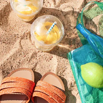 日差しの強い砂浜に並ぶ、夏のリゾートを感じさせるサンダルと冷たいドリンク。