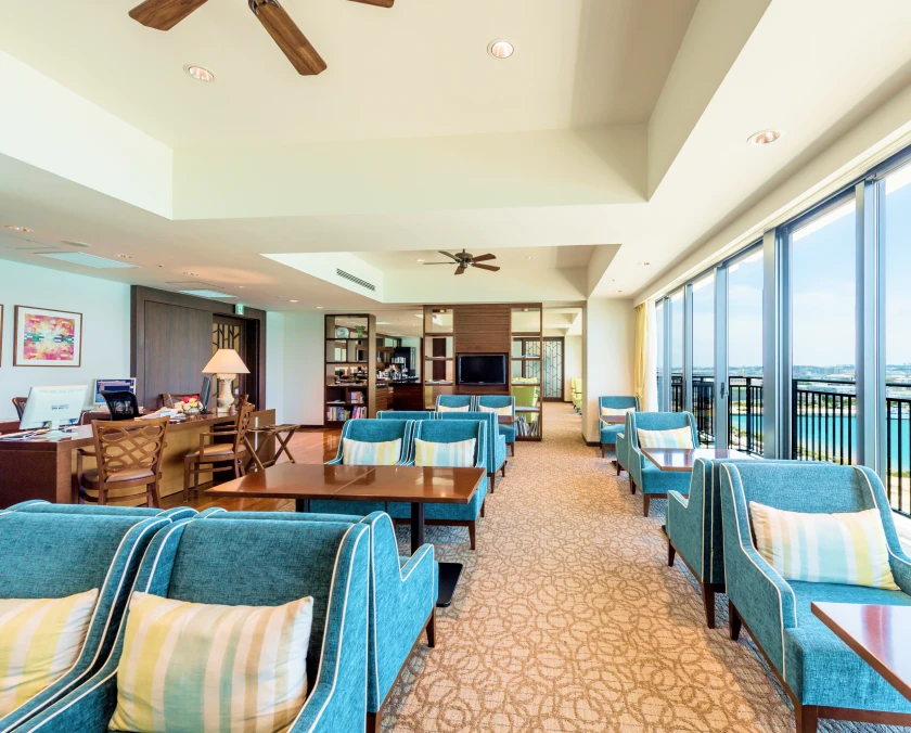 青いソファが並ぶ開放的で上質なラウンジの風景。左側のデスクには笑顔のコンシェルジュが控え、右側の大きな窓からは明るい光が差し込んでいます。