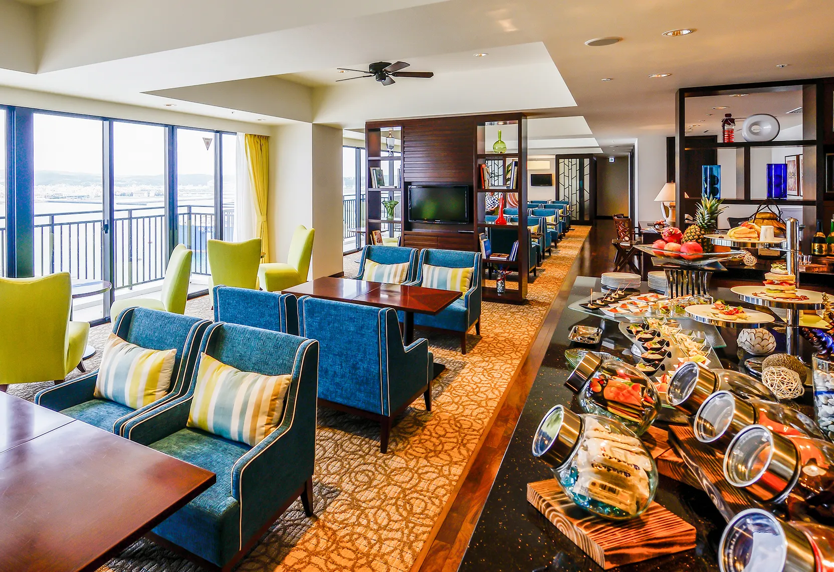 ラウンジの一角に用意された豪華なティータイムのセッティング。手前のカウンターには色とりどりのスイーツや軽食が並び、その奥には海を眺めながらくつろげるソファ席が用意されています。