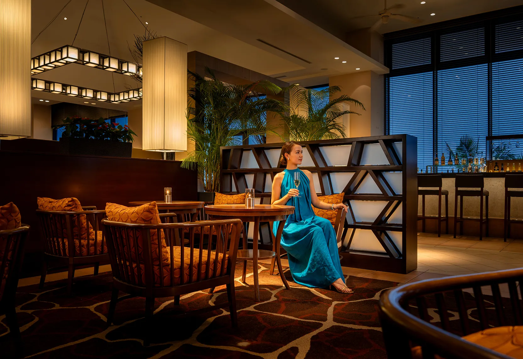 温かい照明に包まれた夜のリゾートレストラン。洗練されたインテリアの中で、青いドレスの女性がシャンパングラスを傾ける優雅なひととき。