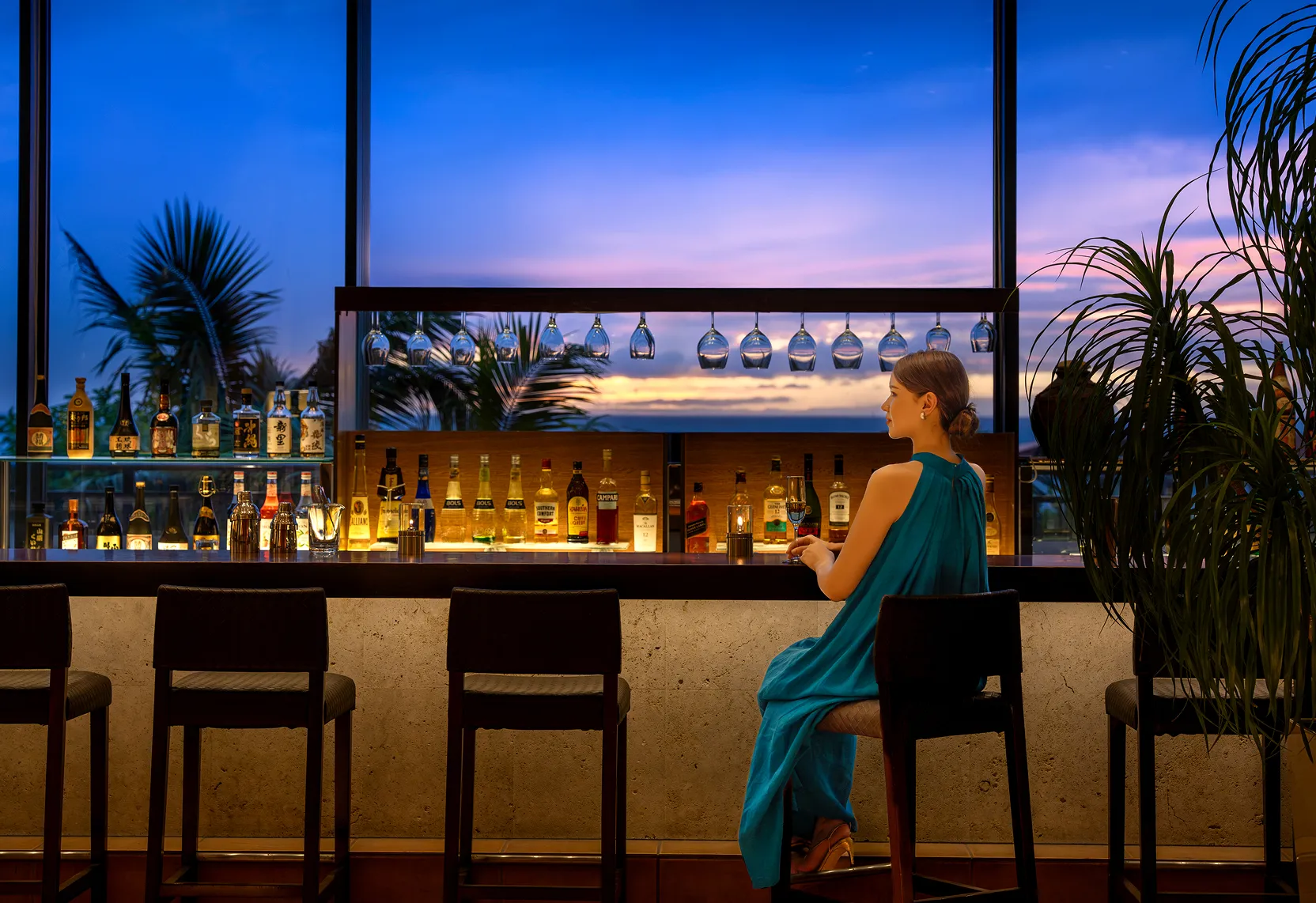 温かみのあるモダンな照明が灯るラウンジ。青いドレスの女性がゆったりとしたソファ席でグラスを傾け、夜のリゾートの空気に浸る様子。
