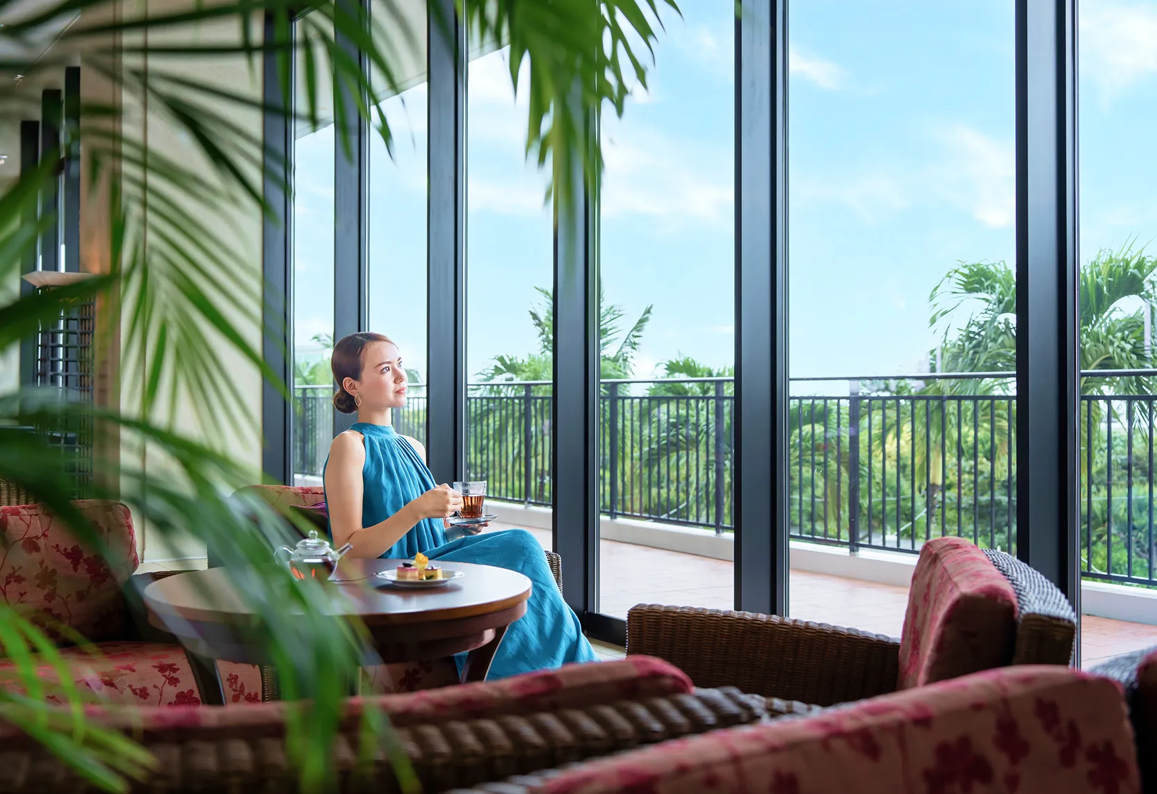 明るい陽光が差し込む開放的なテラス席。青い海と緑を背景に、女性がティータイムを楽しみながら穏やかなひとときを過ごす風景。