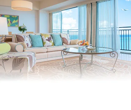 ボタニカル柄のクッションが並ぶ明るいリビング。大きな窓から青い海が一望でき、遠くの空には飛行機がゆったりと飛んでいる景色。