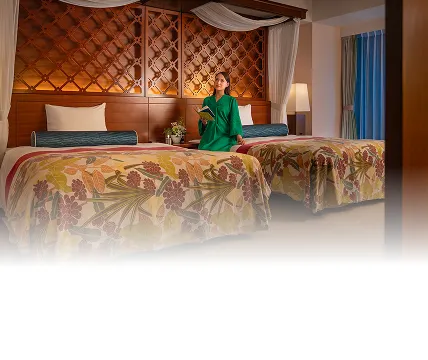 重厚な木彫りのヘッドボードと天蓋が美しいベッドルーム。グリーンのガウンを着た女性がベッドに座り、読書を楽しみながらくつろぐ様子。