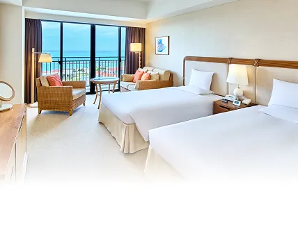 白を基調とした清潔感のあるツインルーム。ラタン調のソファとオレンジのクッションが配され、窓の外には穏やかな海が広がっている様子。